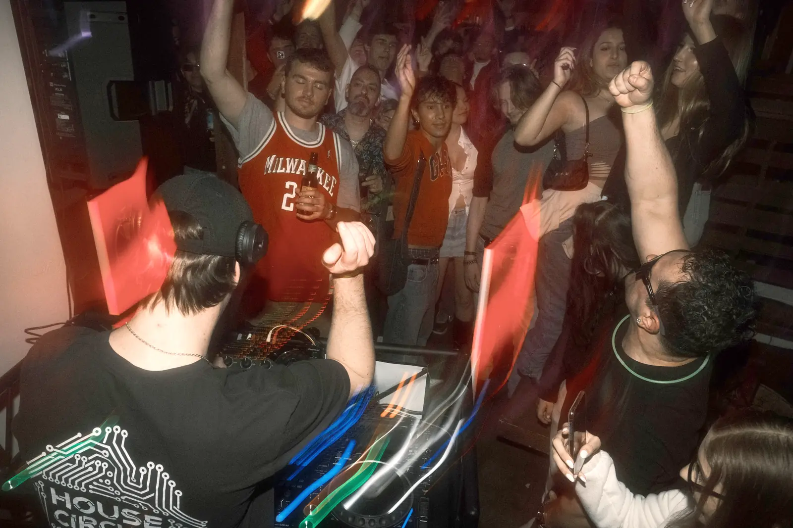 Hands up in front of the DJ, in a House Circuit shirt, on a packed floor